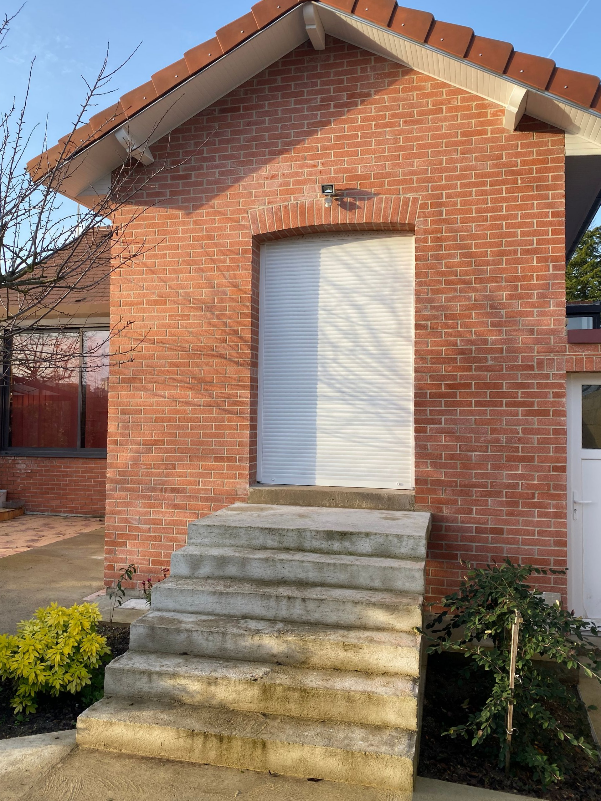 Bâtiment en briques avec marches en béton, volet blanc fermé et toit rouge.