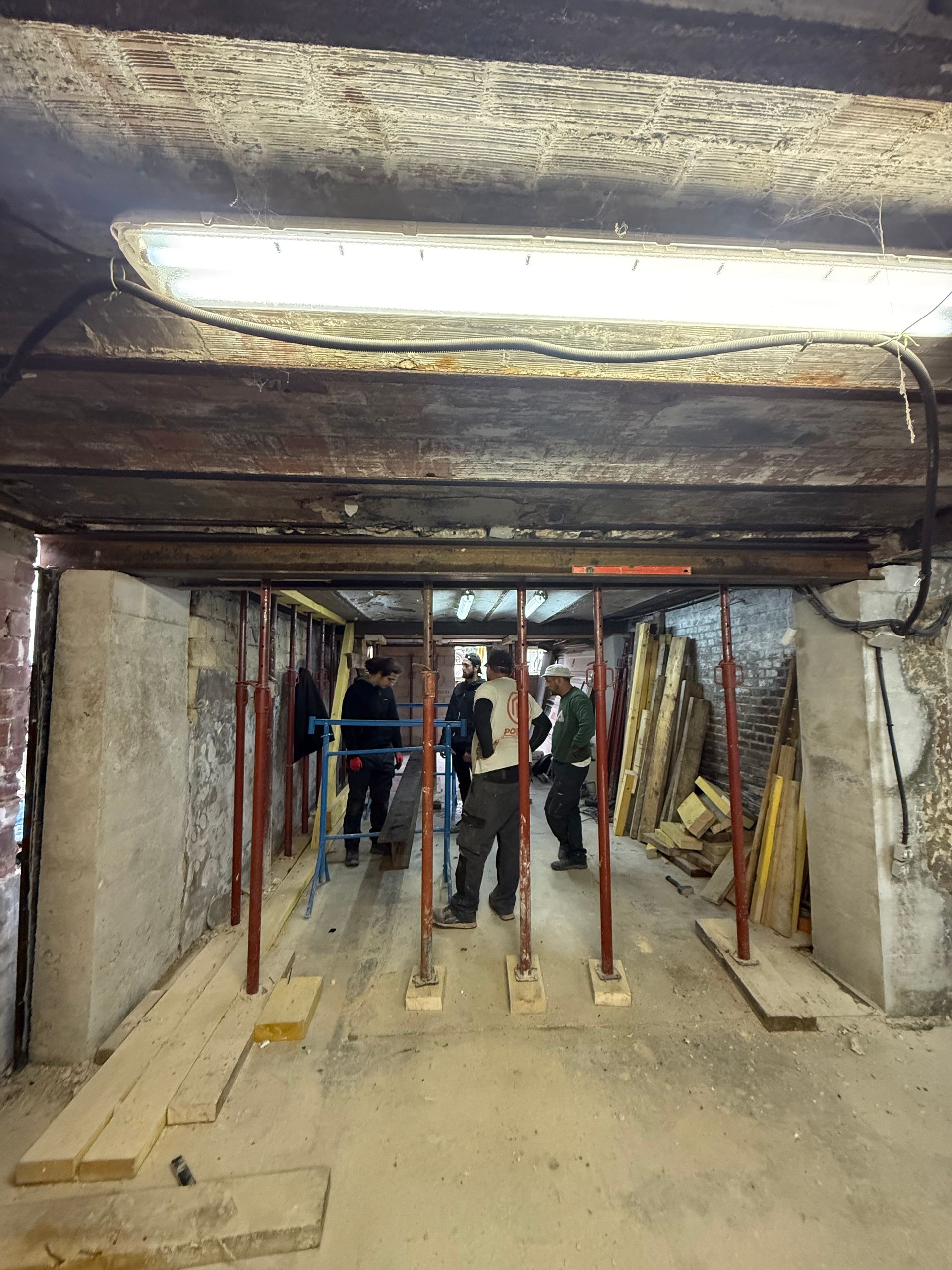 Intérieur d'un chantier de construction ; poutres en bois, supports métalliques et ouvriers en arrière-plan.