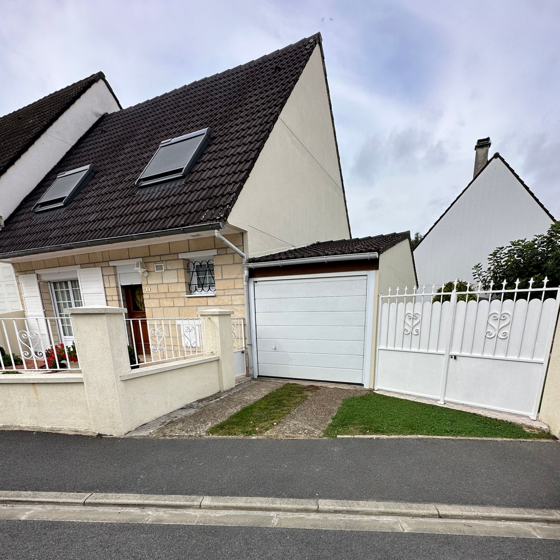 Maison avec un toit foncé, une porte de garage blanche, une petite clôture et une allée.