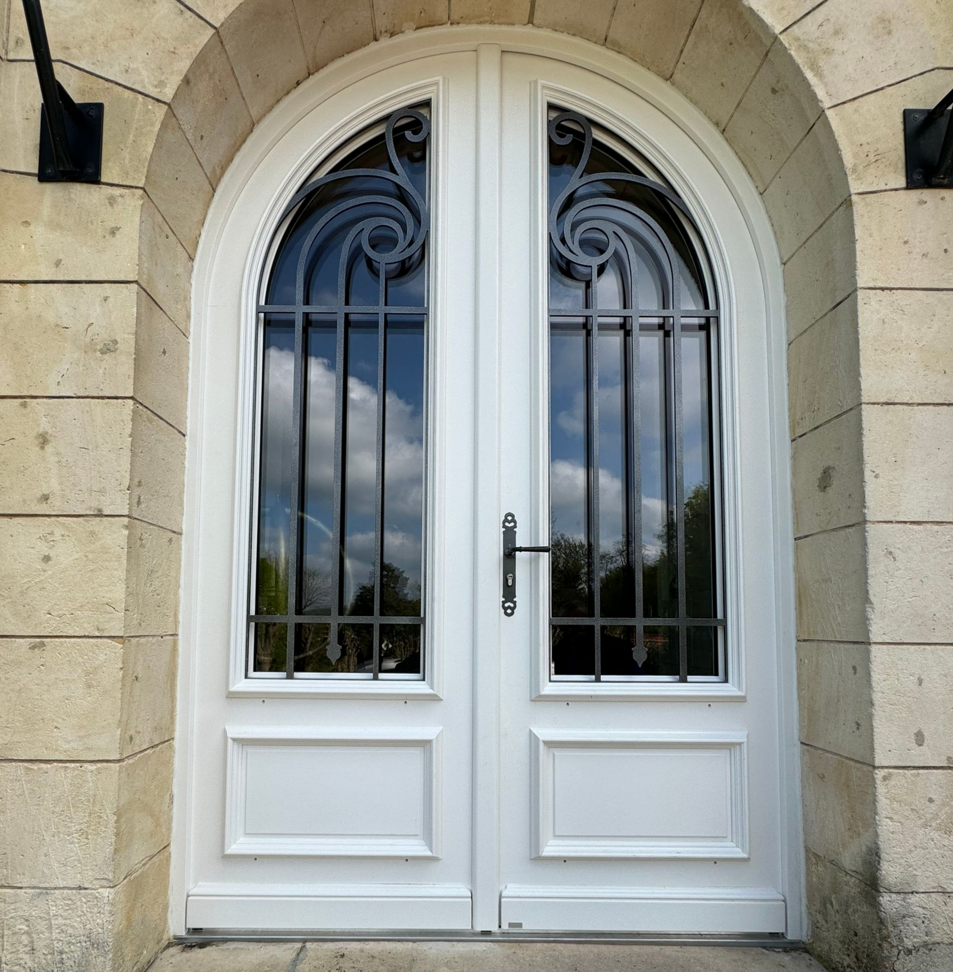 Double porte blanche, ferronnerie noire, encadrée par une arche en pierre.