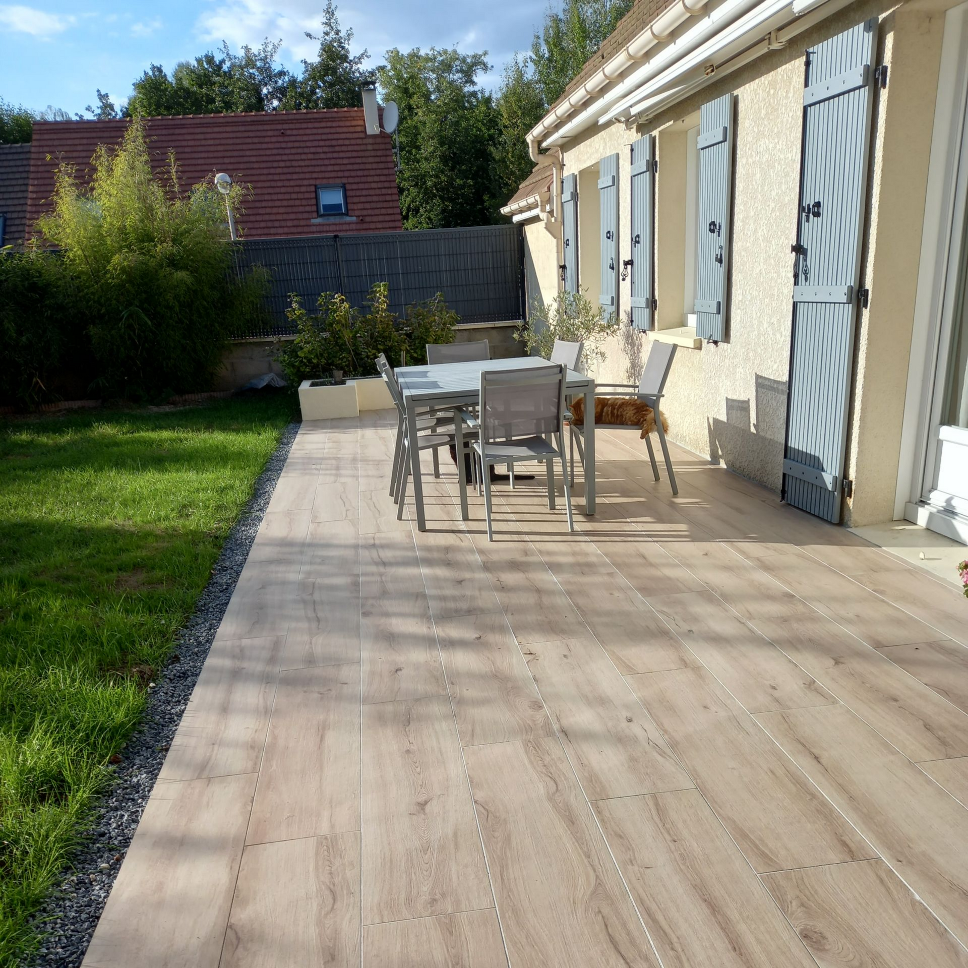 Terrasse avec table à manger extérieure, à côté d'une maison aux volets bleus.