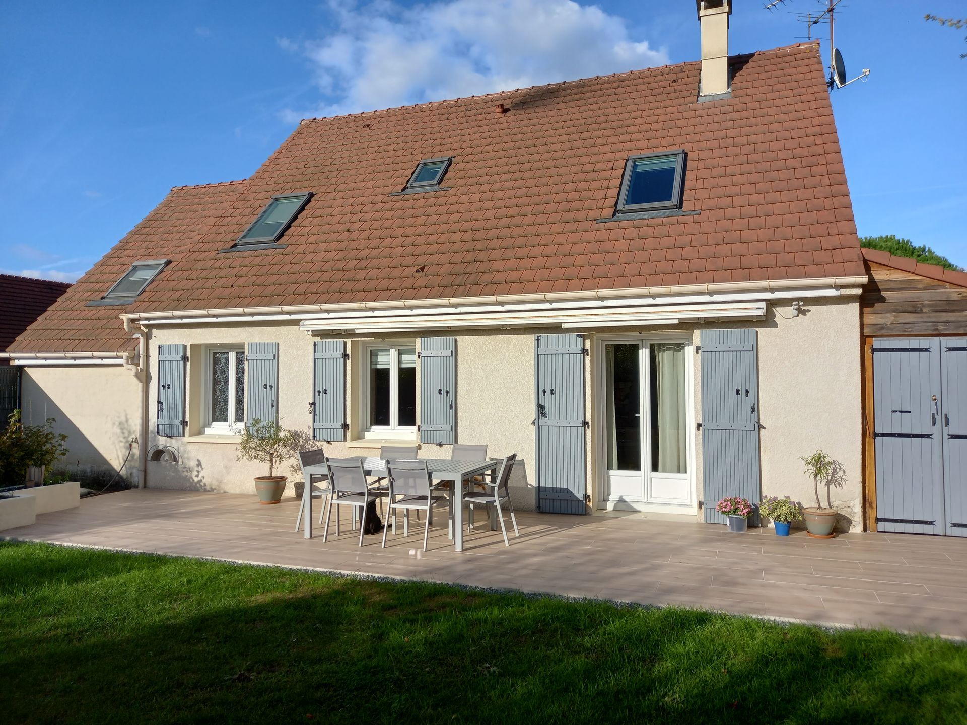 Maison avec toit en tuiles, volets bleus et mobilier de terrasse.