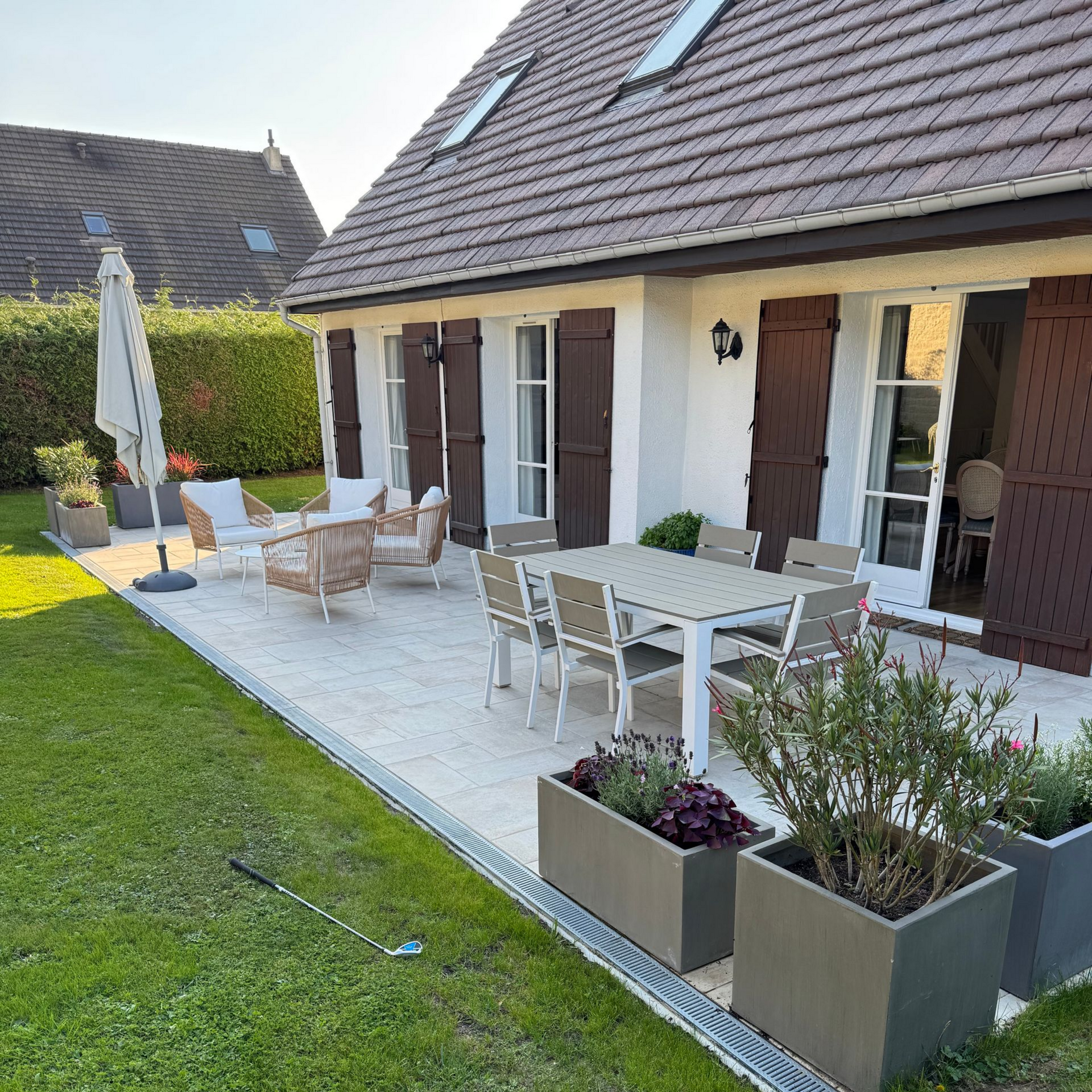 Terrasse avec ensemble de repas extérieur et coin salon près d'une maison blanche aux volets marron.