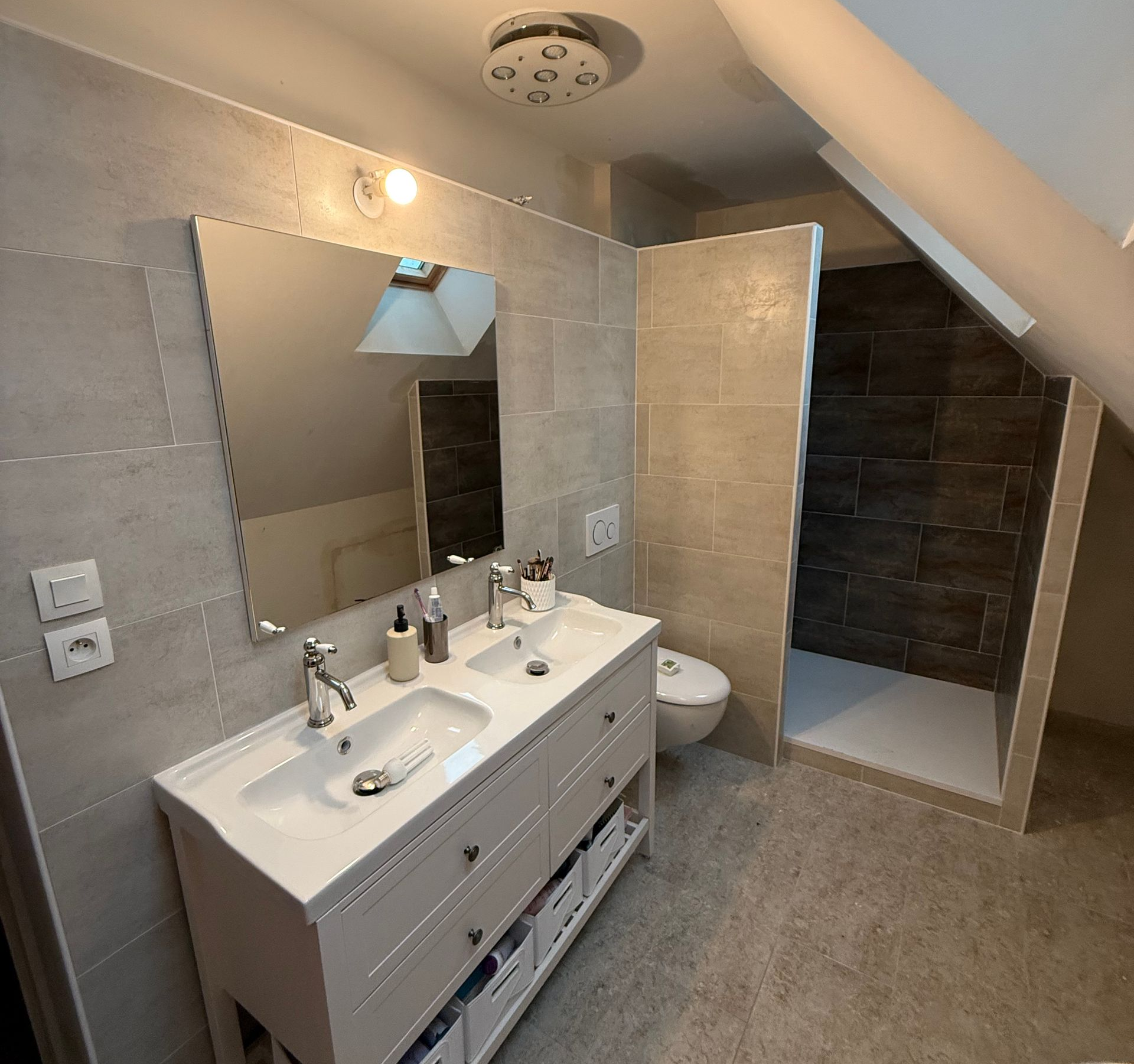 Salle de bains avec double vasque blanche, grand miroir, douche à l'italienne et plafond en pente.