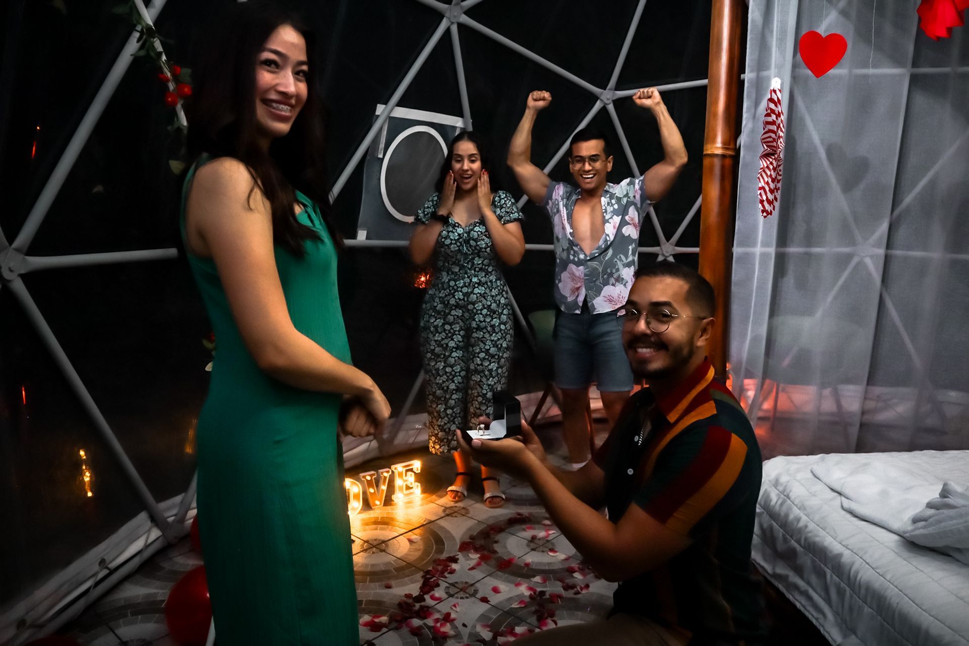 Man kneeling, proposing to woman in a dome. Friends cheer in the background, a romantic setting.