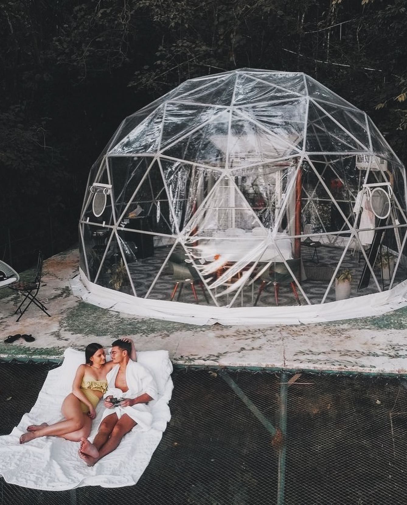 Couple in swimsuits and robes on a netted lounge, kissing. Geodesic dome behind them, outdoor setting.