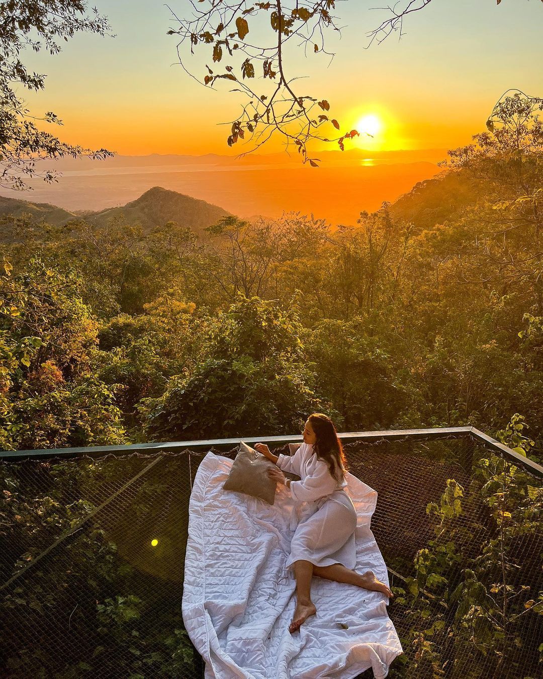 Woman relaxing on a net bed, enjoying a sunset view over a forest.