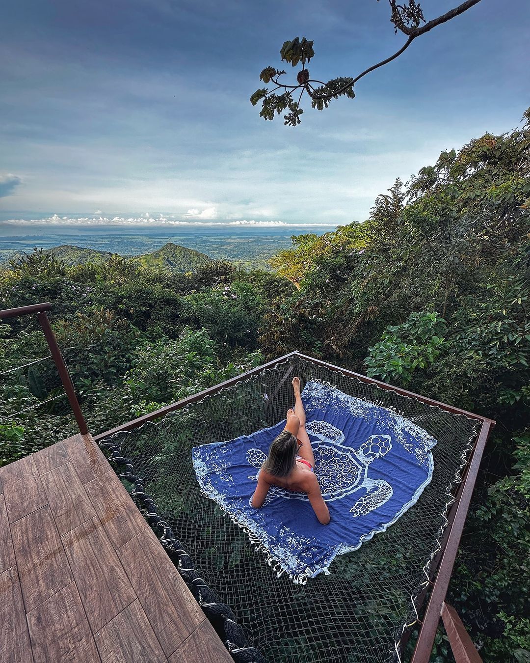Woman relaxes on a net platform overlooking a lush landscape, blue blanket, cloudy sky.