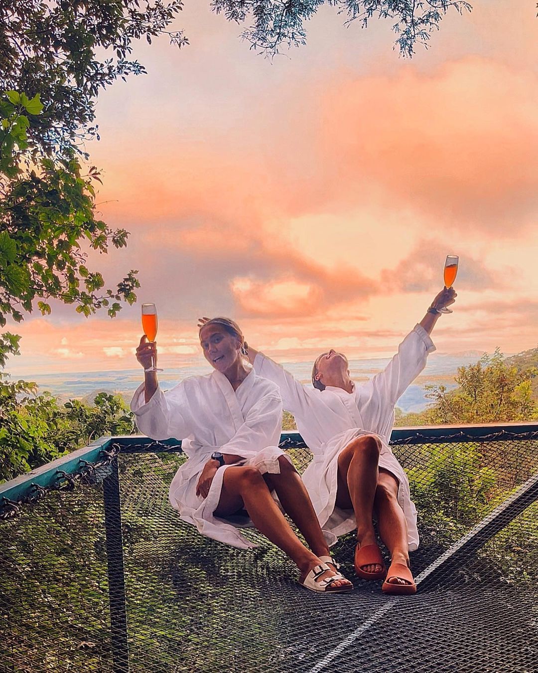 Two people in white robes raise drinks, enjoying a sunset view from a net.