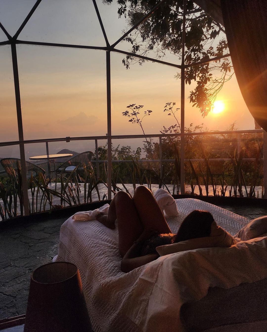 Person relaxing on a bed, watching the sunset over a landscape.