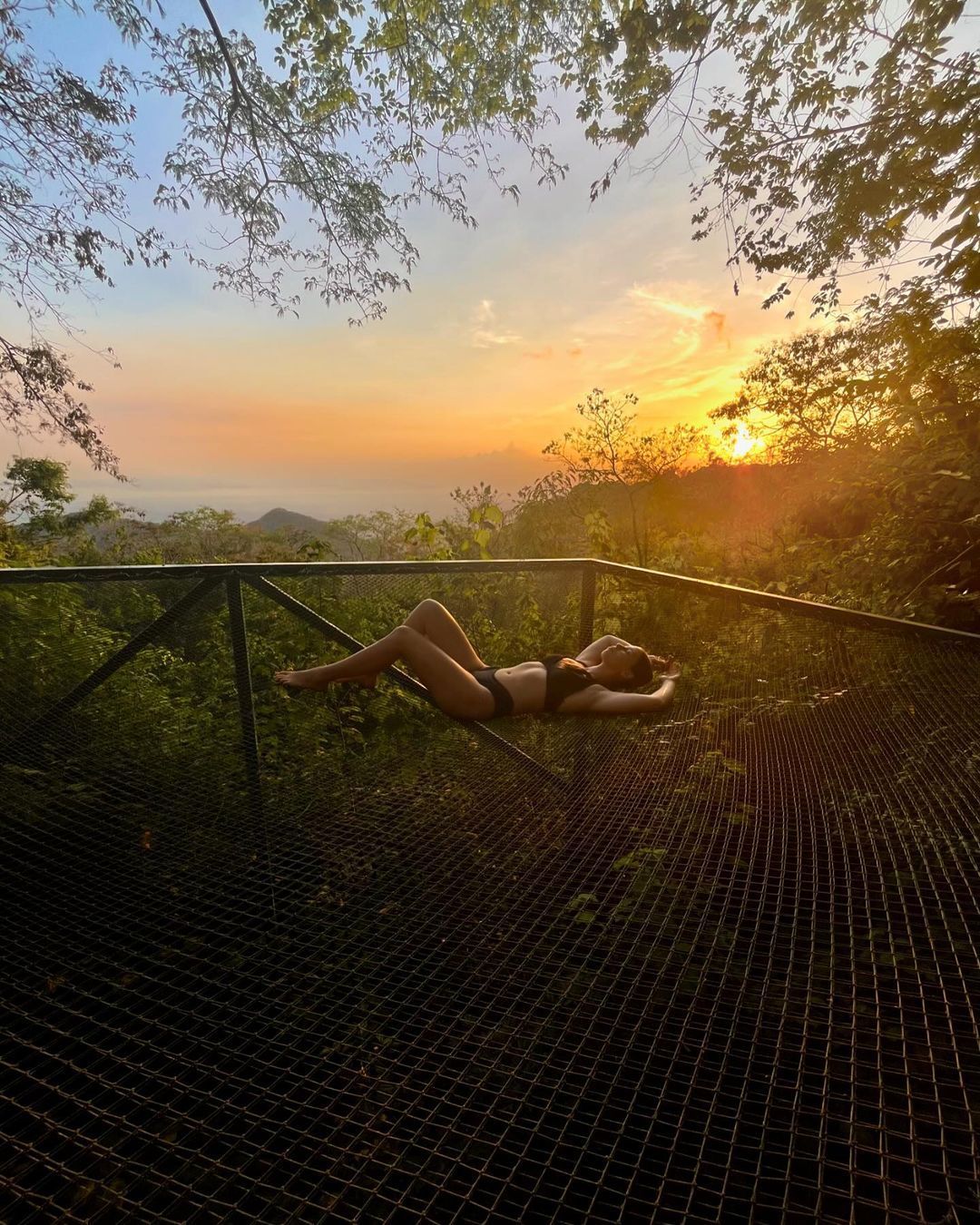 Person reclines in a hammock at sunset, overlooking a landscape with lush greenery, blue sky, and golden sunlight.