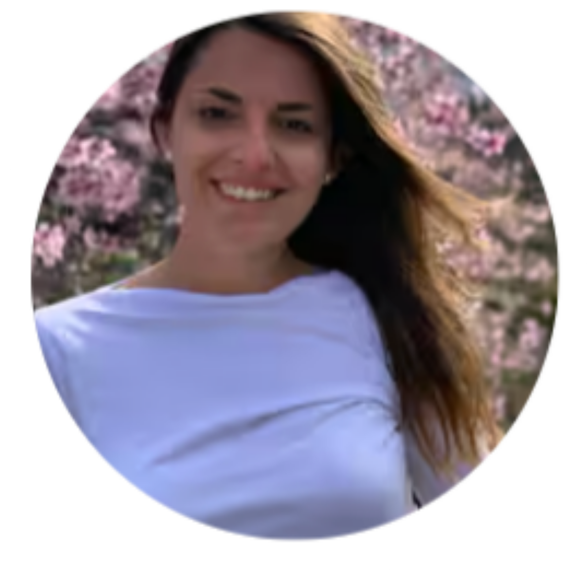 Woman smiles in front of pink flowering trees. Wearing a white top, brown hair.
