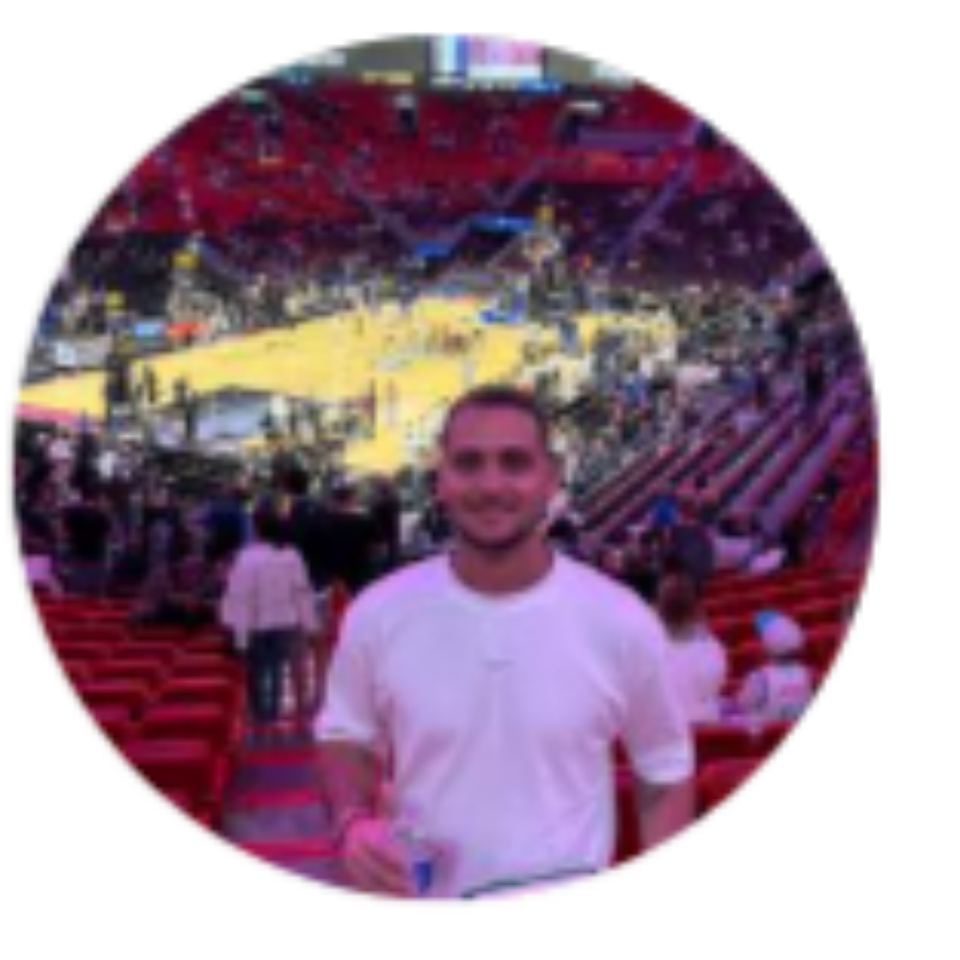 Man in white shirt smiling at a basketball game. Red stadium seats and court in background.