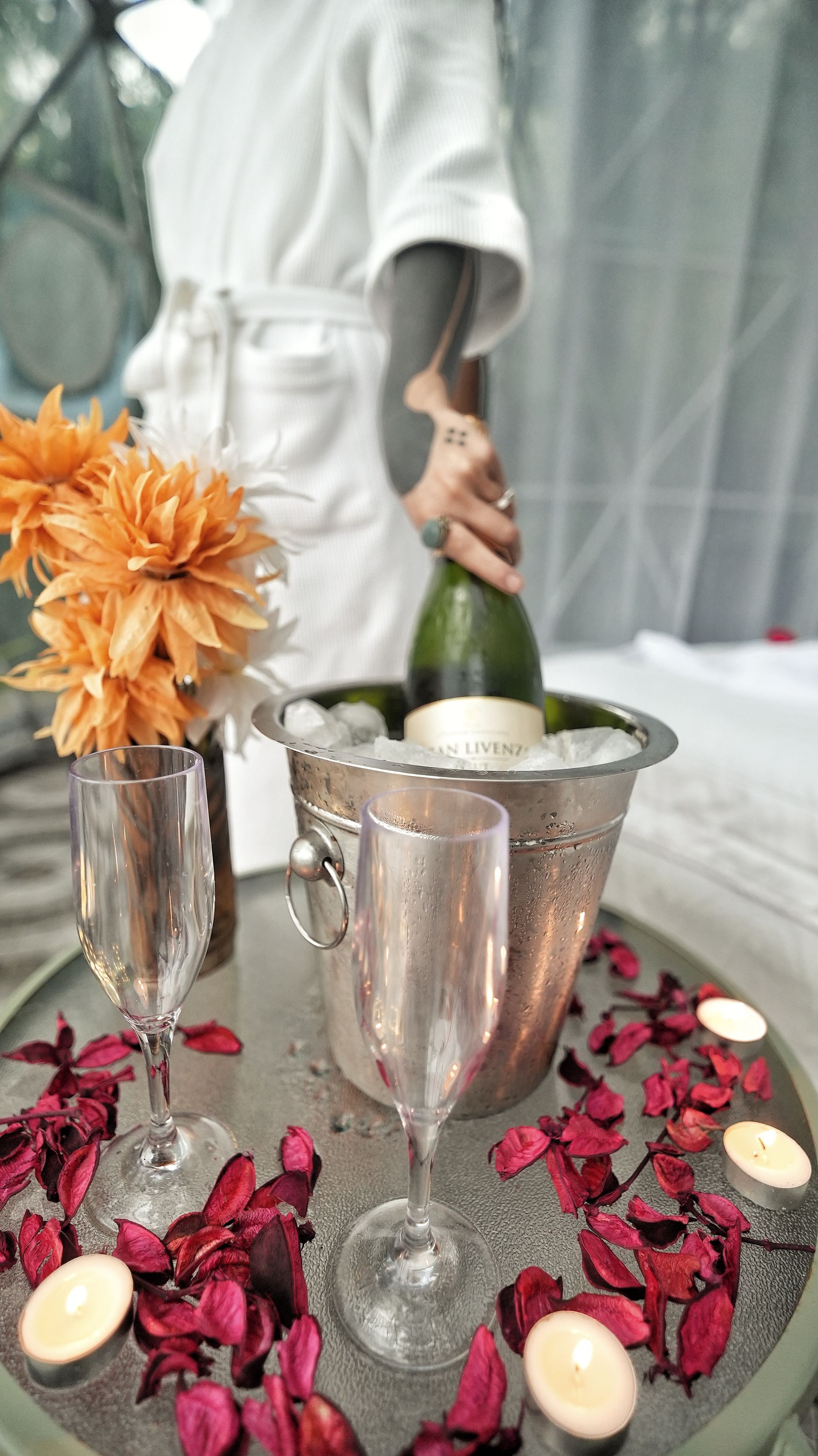 Person in white robe opening champagne bottle in a silver ice bucket, flutes and candles on a table with flowers and petals.