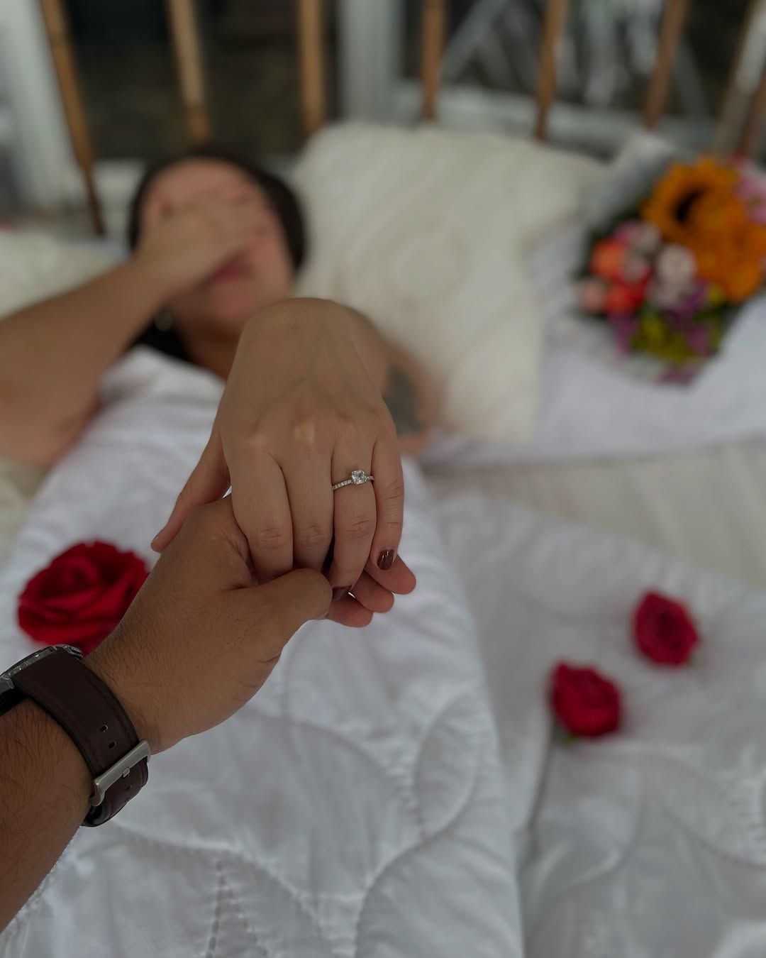 Person's hand with engagement ring held by another's. Person lying in bed, eyes covered. Flowers and rose petals nearby.