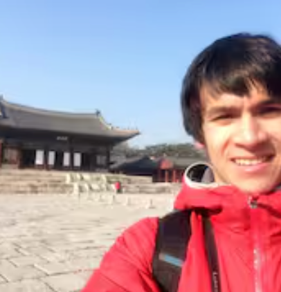 A man in a red jacket is taking a selfie in front of a building.