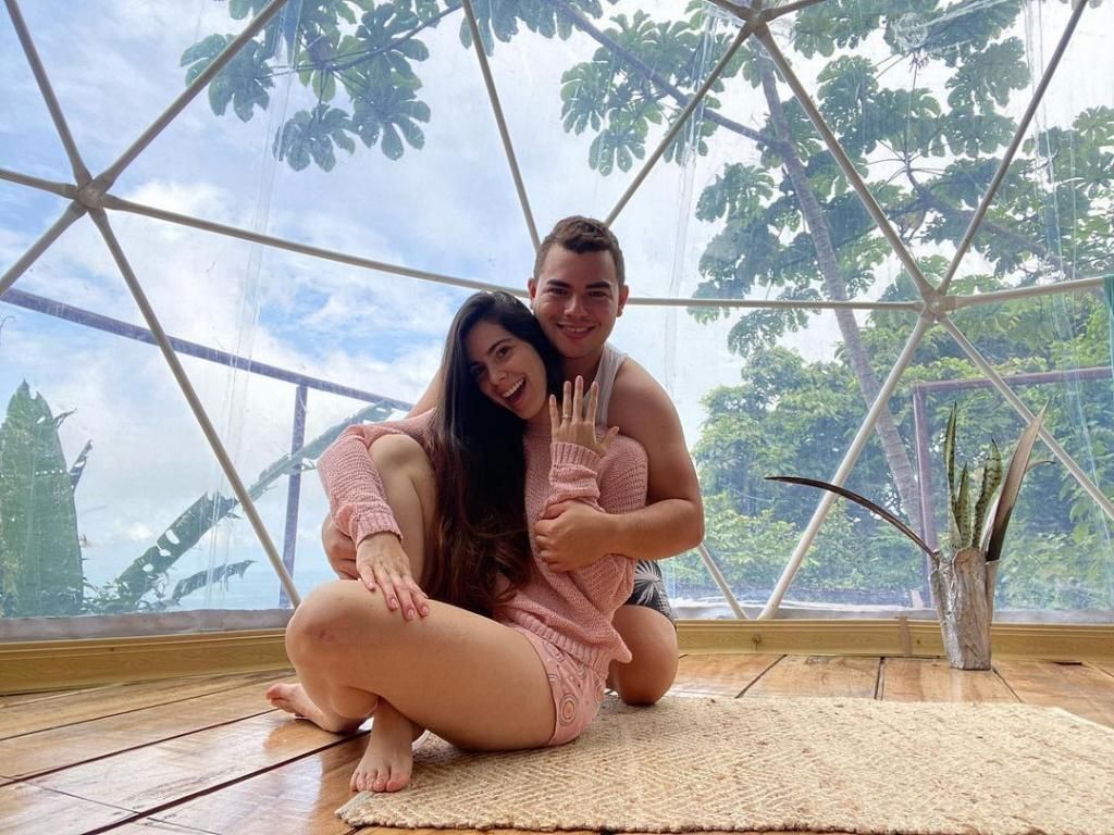 Pareja sonriendo y abrazándose dentro de una cúpula geodésica con vista a árboles verdes.