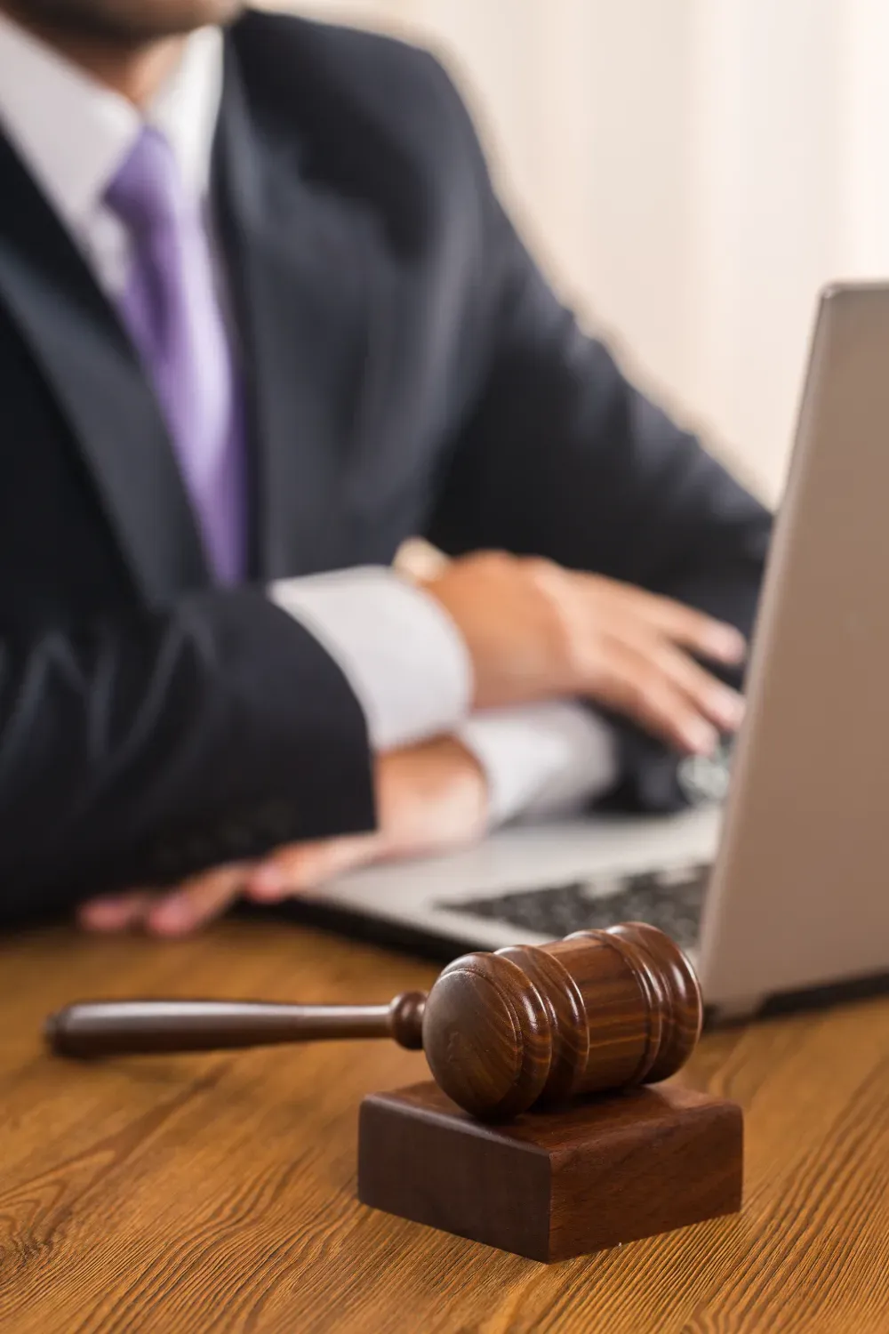 Una persona con traje y corbata violeta está sentada en un escritorio usando una computadora portátil, con un mazo de juez