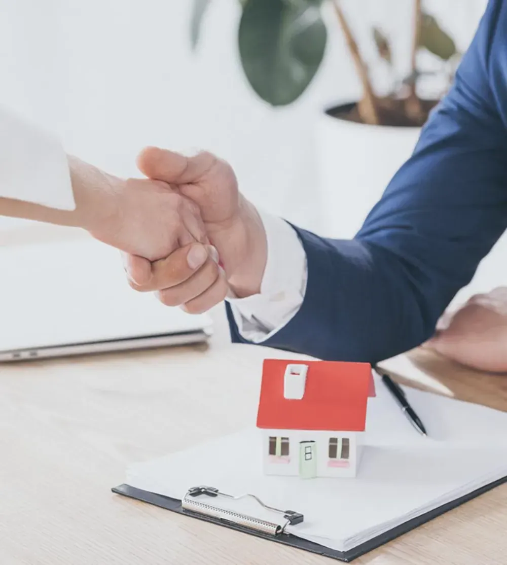Dos personas vestidas de manera profesional se dan la mano mientras firman un contrato con una pequeña maqueta de casa 
