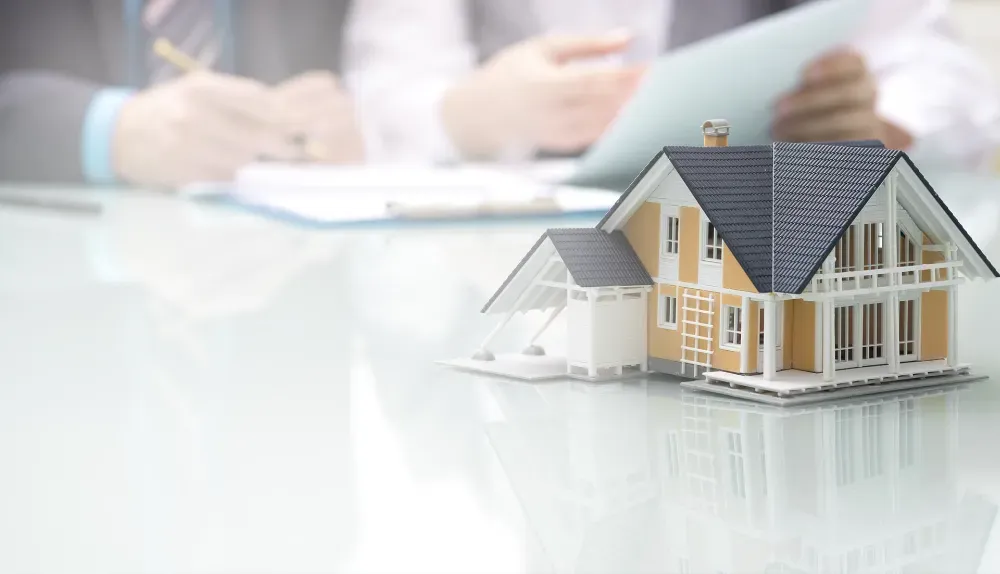 Una maqueta de una casa suburbana reposa sobre una mesa de cristal frente a personas que revisan documentos en una oficina.