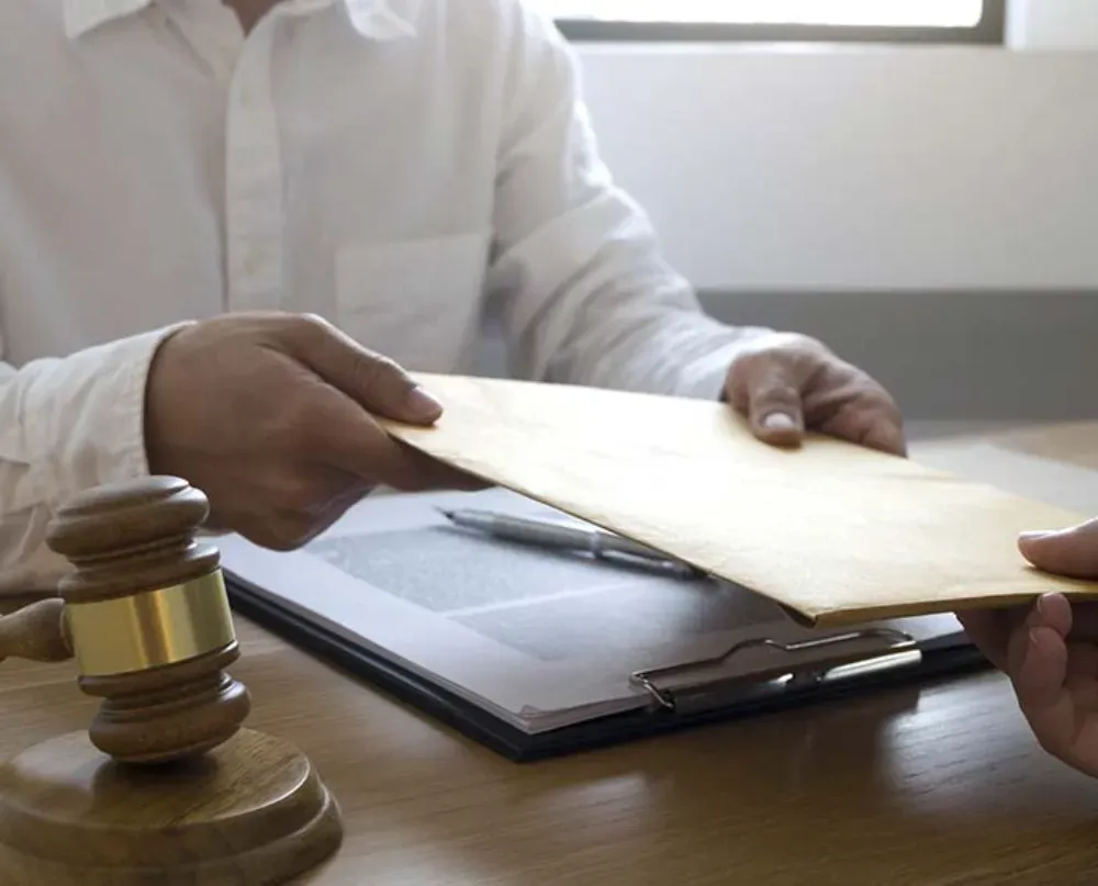 Una persona con una camisa blanca le entrega un sobre marrón a otra persona sobre un escritorio con un mazo de madera
