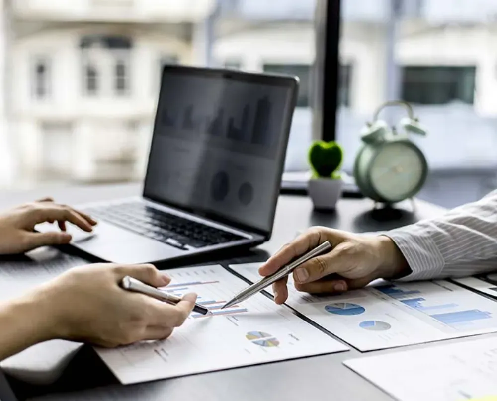 Dos personas revisando gráficos y datos en papeles en un escritorio con una computadora portátil