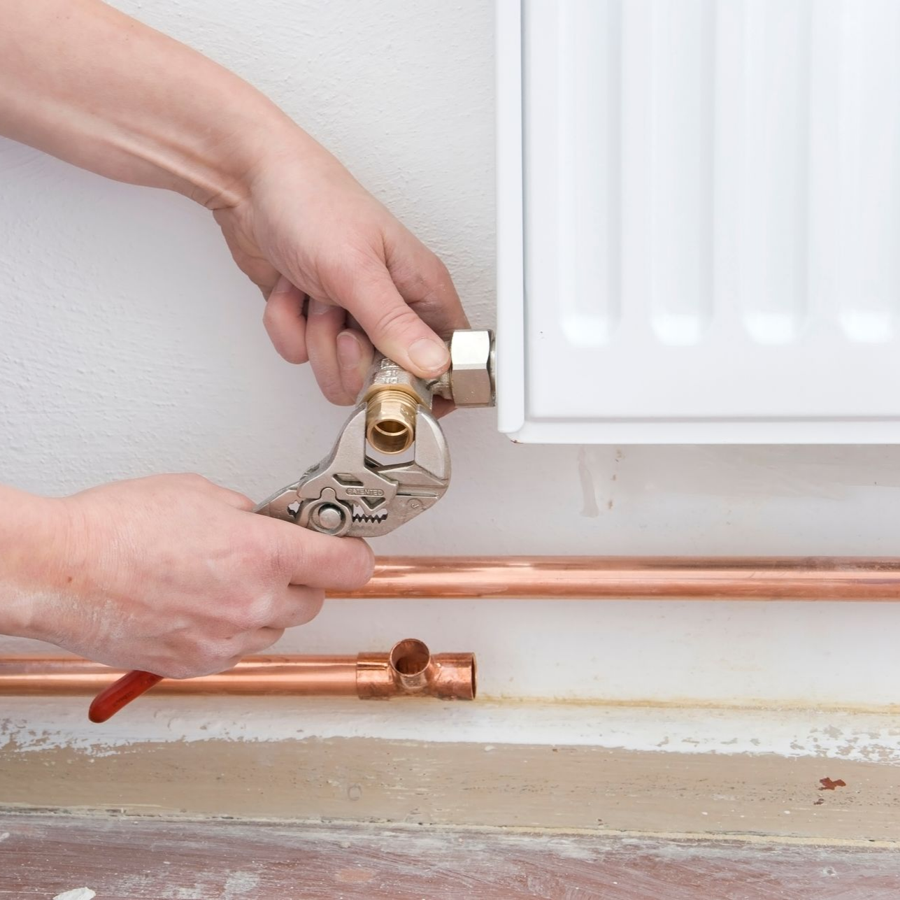 Des mains utilisent une clé pour serrer un raccord sur un radiateur près de tuyaux en cuivre.
