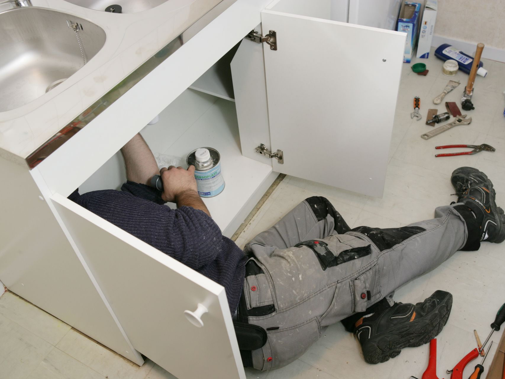 Une personne travaillant sous un évier de cuisine, entourée d'outils et de fournitures.