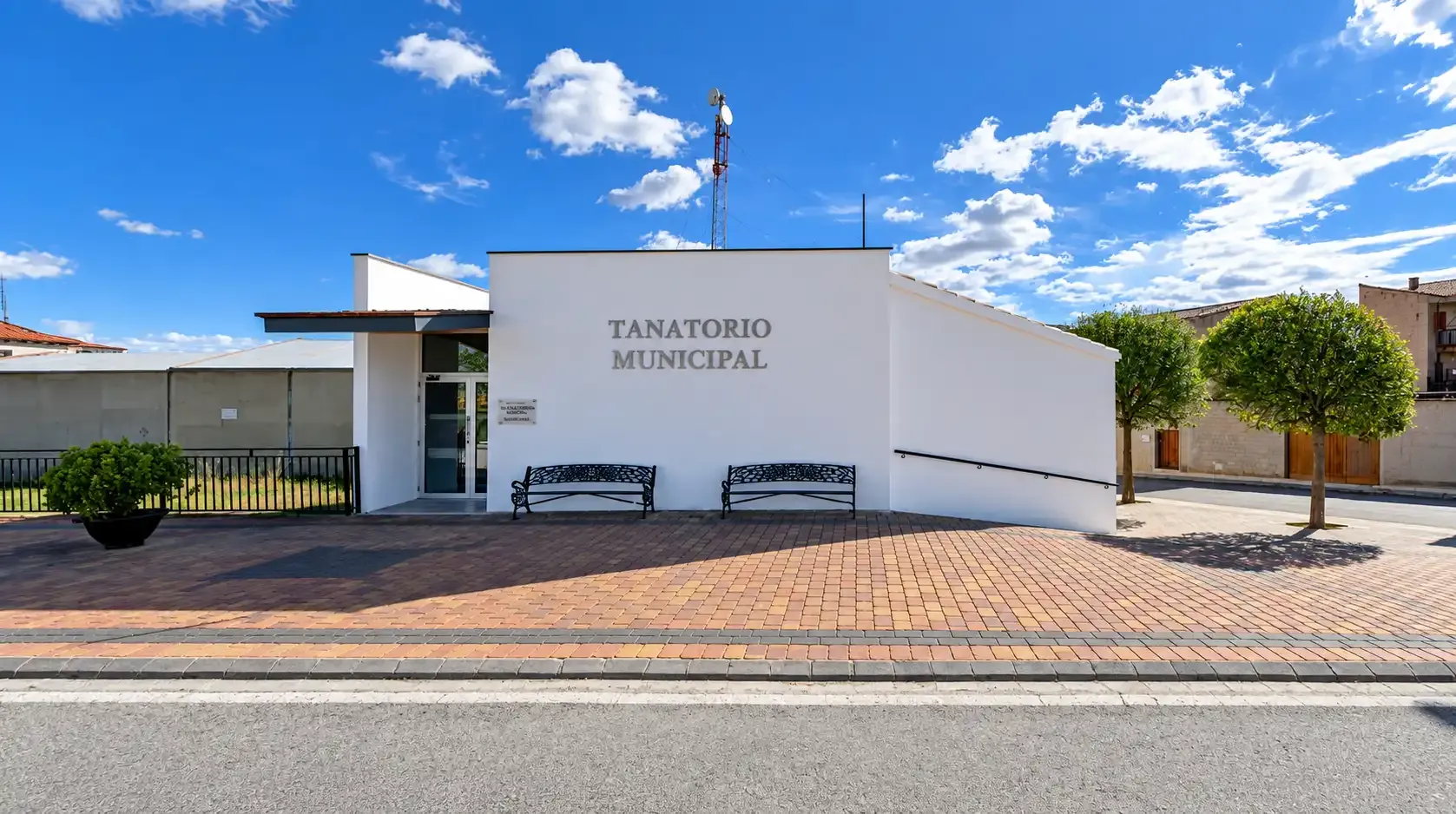 Edificio blanco moderno con el rótulo «TEACHERS HOME», bancos y un jardín delantero de estilo desértico bajo un cielo azul.