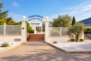 Entrada al TANATORIO con escalones cerrados por una verja, muros de piedra y árboles bajo un cielo despejado