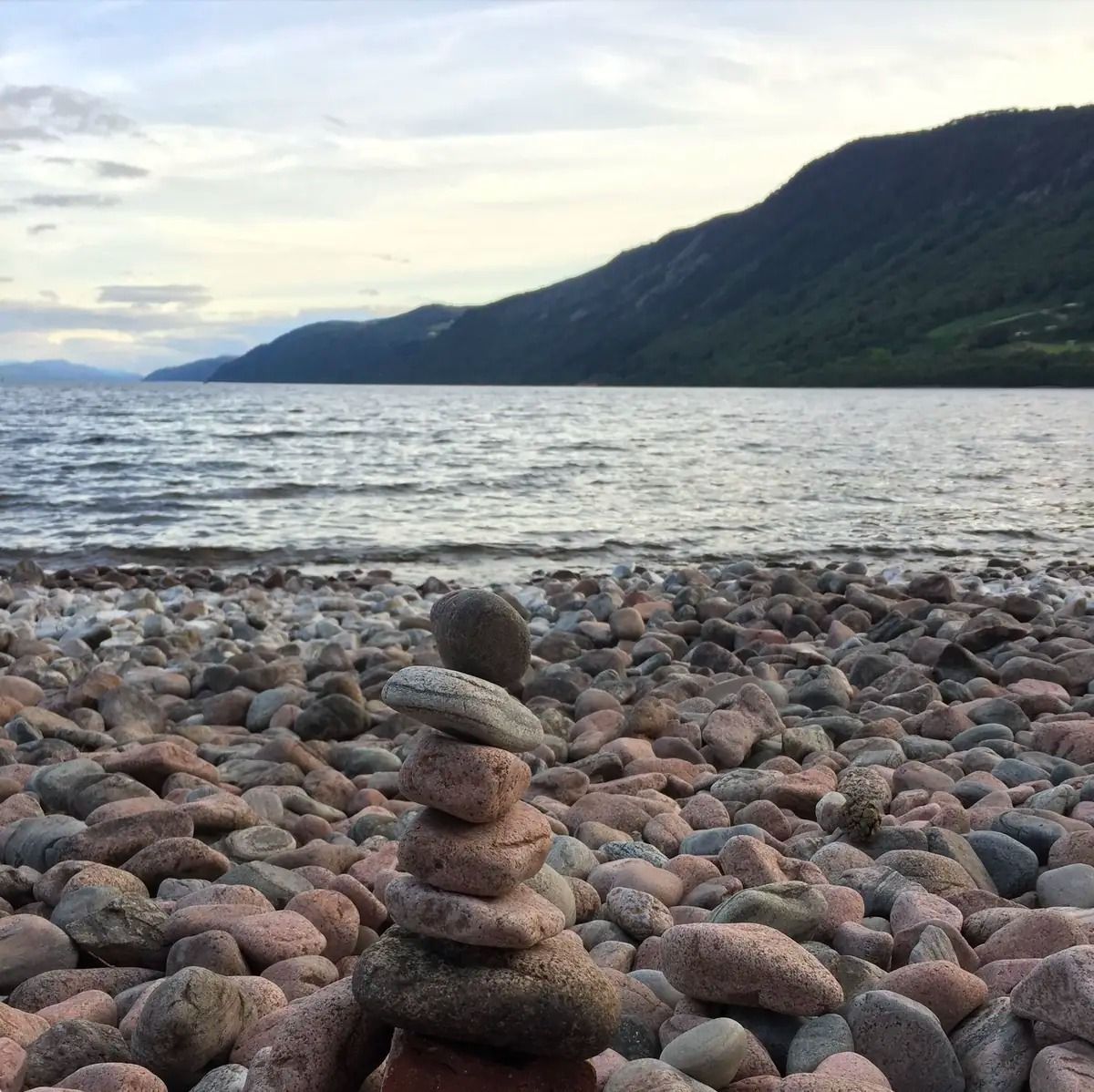 Piedras apiladas en una playa rocosa con un lago tranquilo y montañas boscosas al atardecer.