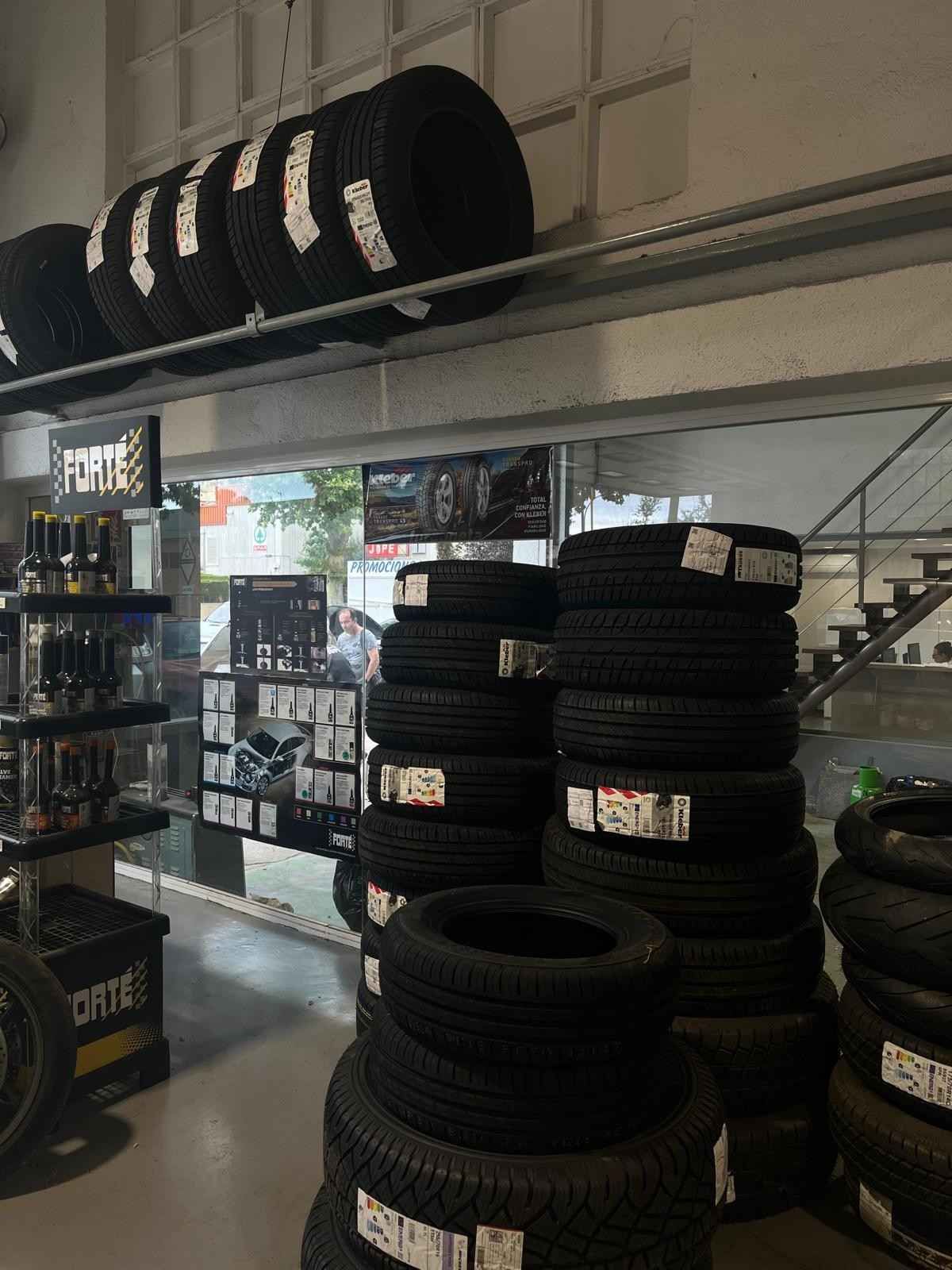 Un mecánico con un mono azul mira la parte inferior de un coche en un taller, sosteniendo una herramienta metálica.