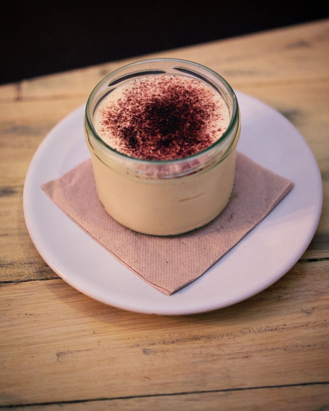 Postre de cristal espolvoreado con cacao en polvo sobre una servilleta encima de un plato blanco sobre una mesa de madera.
