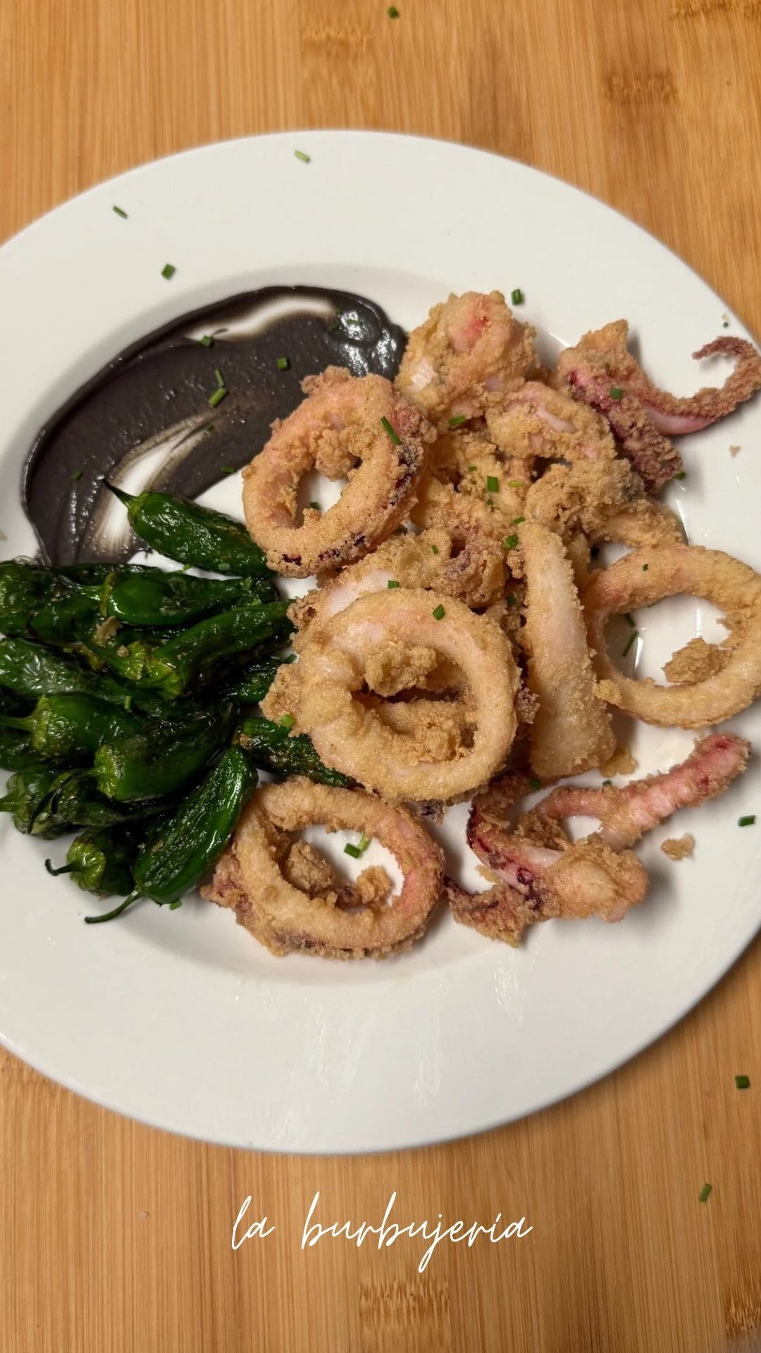 Anillos de calamar a la parrilla en un plato blanco con salsa oscura y verduras salteadas, servidos en una mesa de madera.