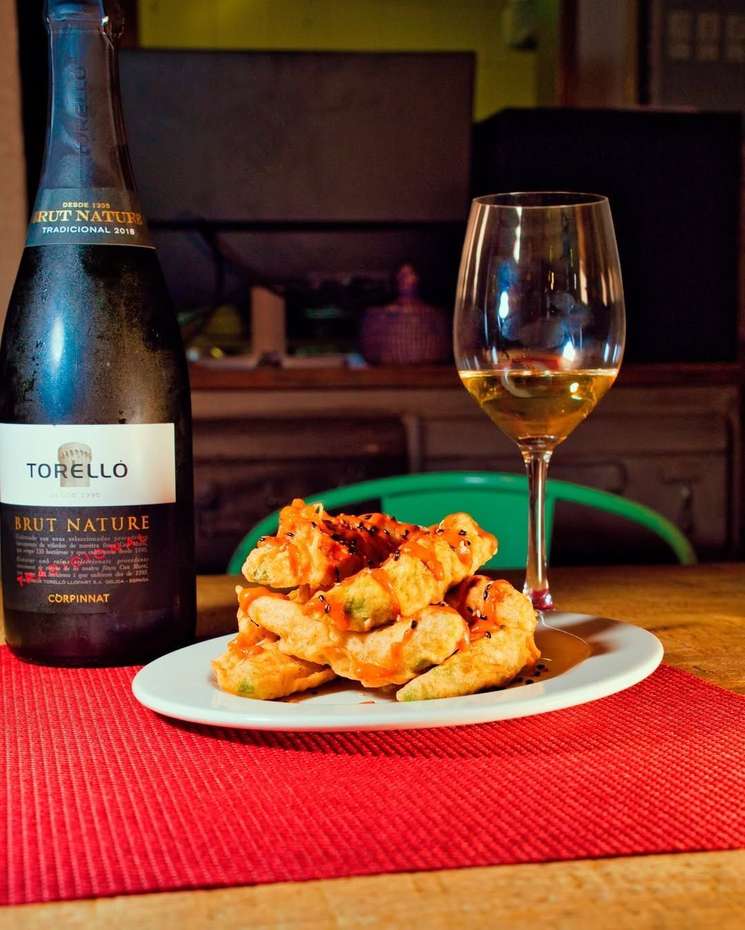 Botella de vino y copa junto a un plato de bocadillos fritos sobre un mantel individual rojo.