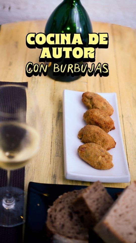 Bebida espumosa embotellada con tapas en un plato blanco y pan en una mesa, con el texto “Cocina de Autor con Burbujas”.