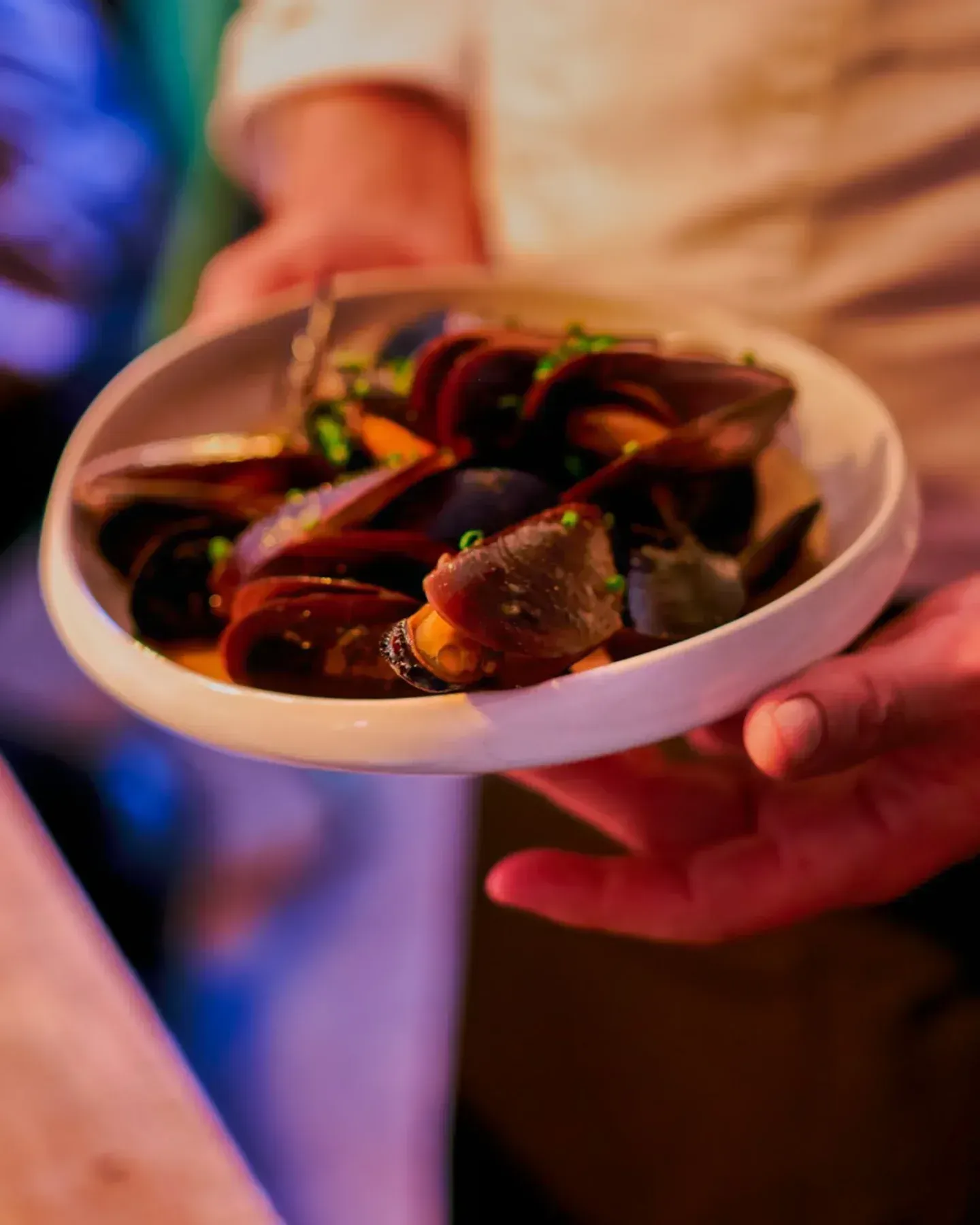 Manos sosteniendo un cuenco de mejillones en una rica salsa oscura, adornados con hierbas, en la mesa de un restaurante.