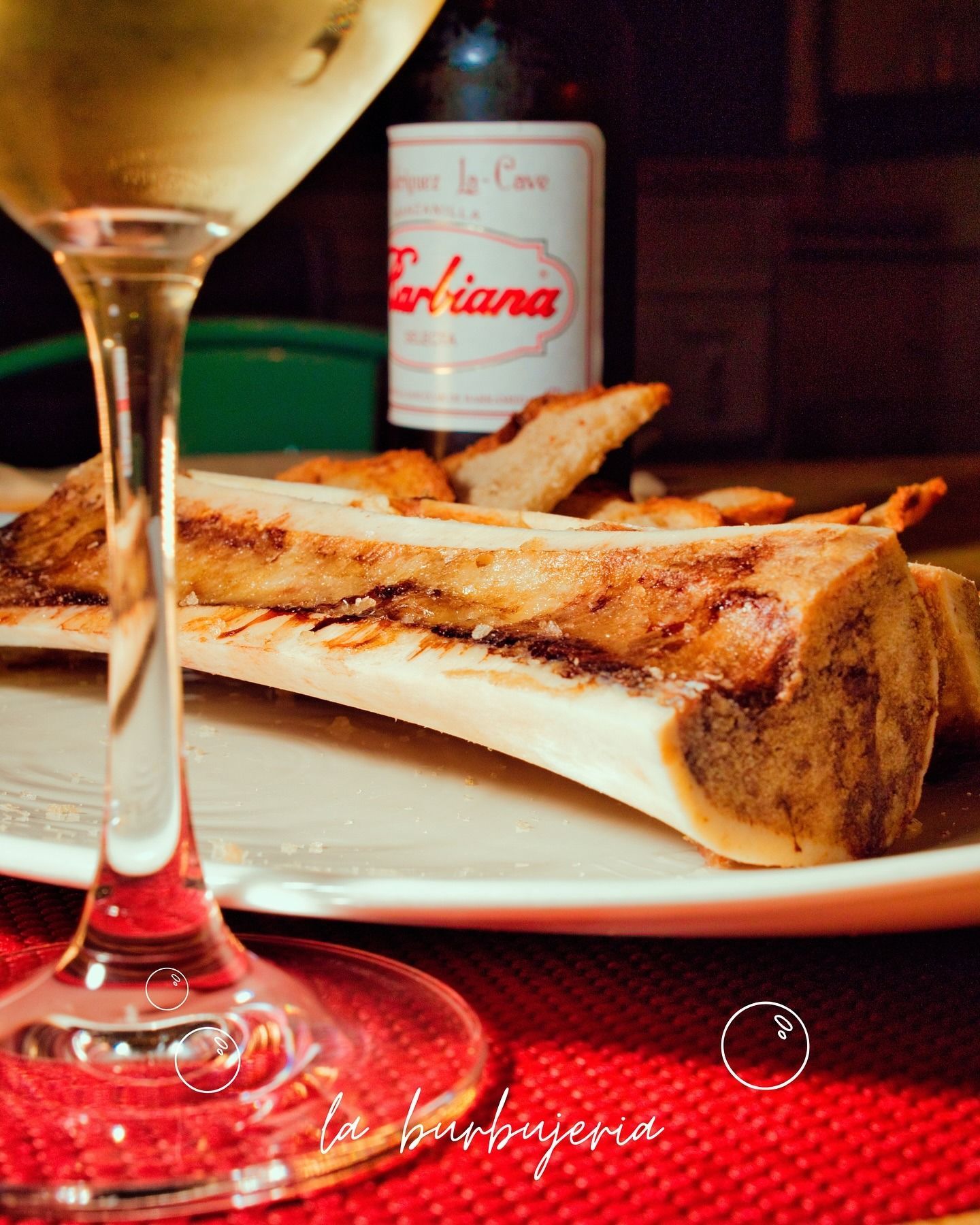 Una copa de vino y una botella junto a un plato de médula ósea sobre un mantel rojo.