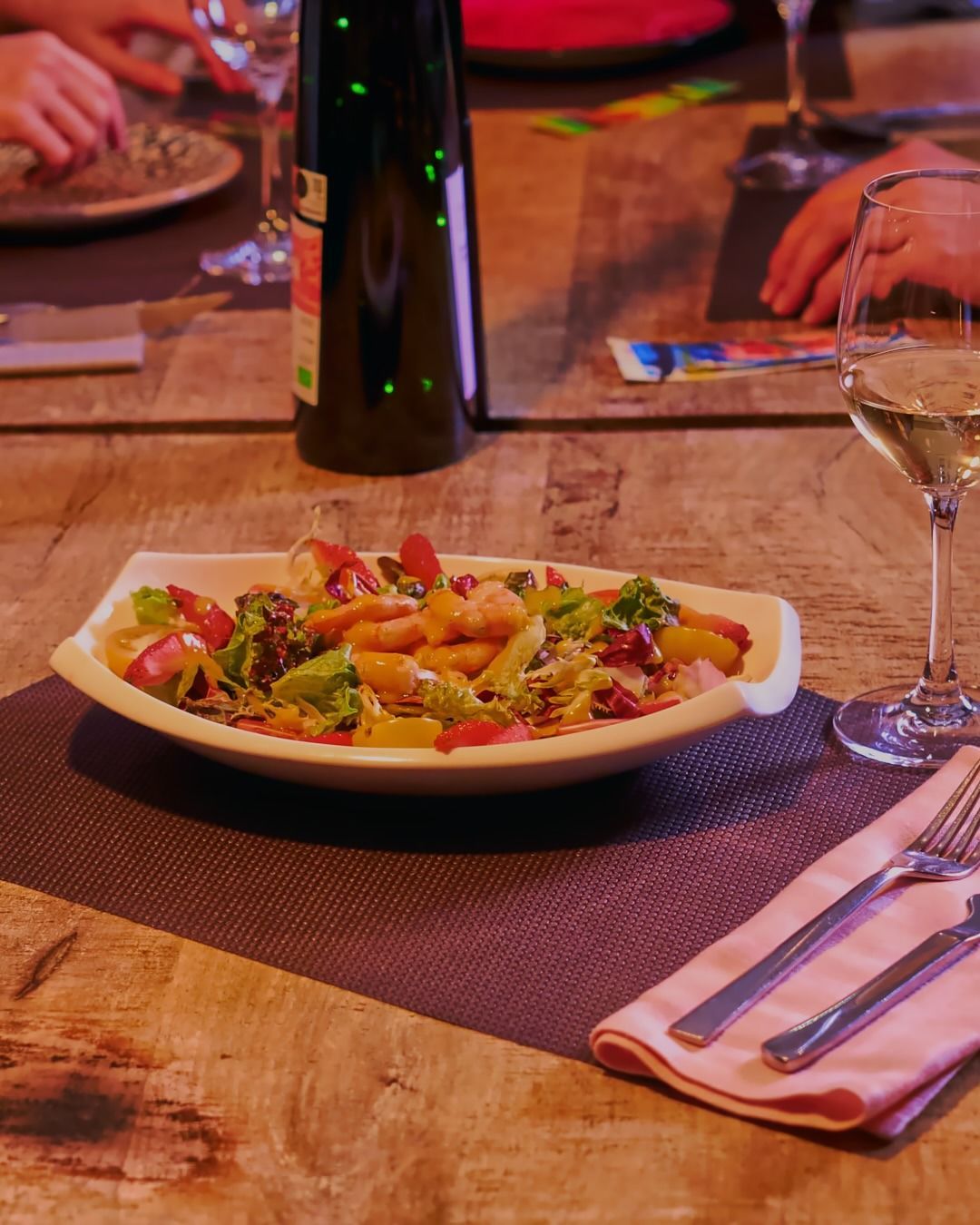 Ensalada servida en un plato sobre una mesa de restaurante con una copa de vino, cubiertos y una iluminación ambiental tenue.