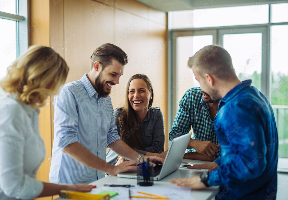 Un groupe de personnes rient, rassemblées autour d'un ordinateur portable dans un bureau moderne, regardant l'écran.
