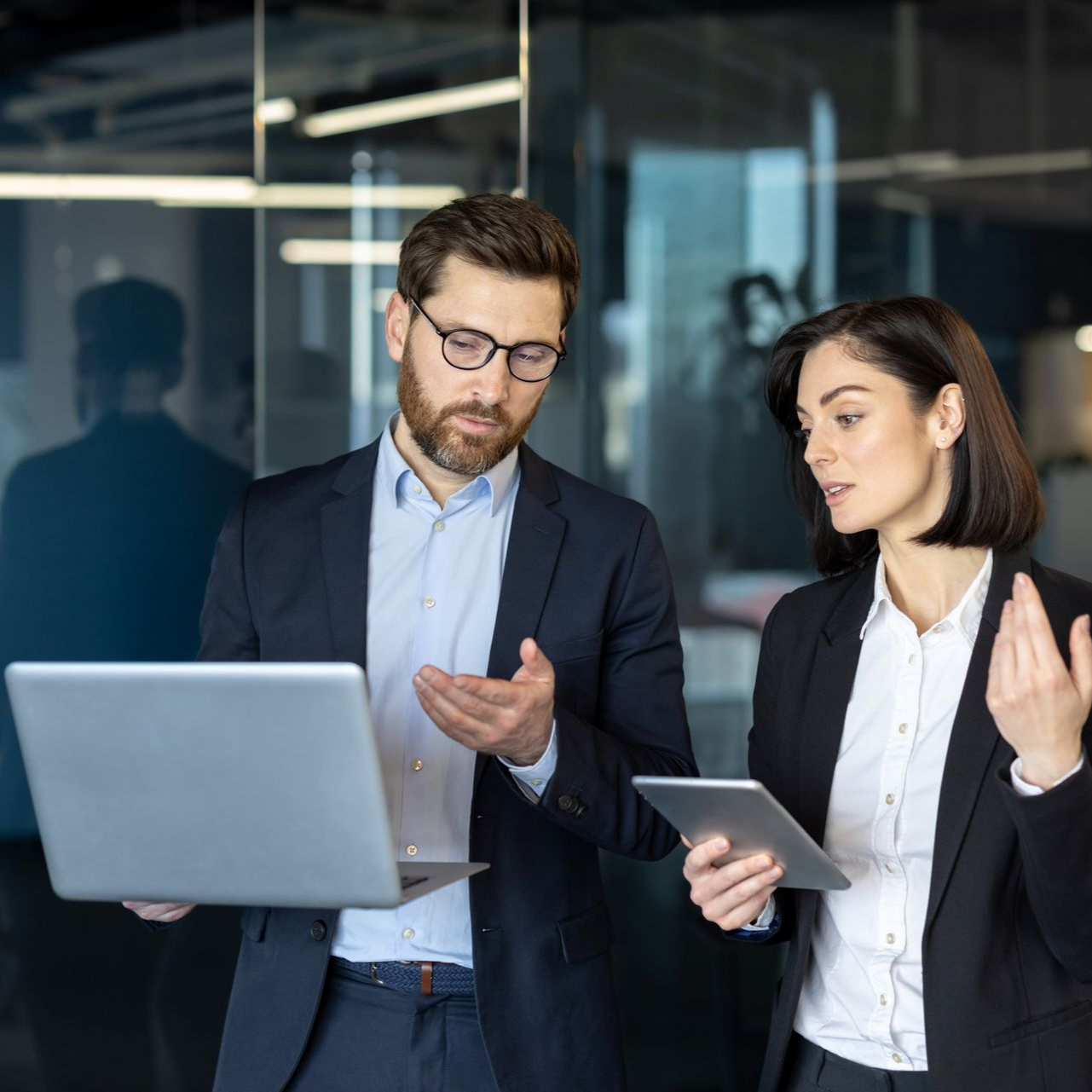 Un homme portant des lunettes et une femme munie d'une tablette discutent sur un ordinateur portable dans un bureau.