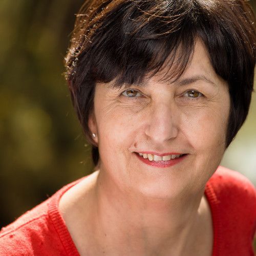 Une femme en pull rouge sourit à l'extérieur, avec de la verdure floue en arrière-plan.