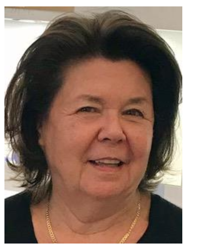 Une femme aux cheveux bruns courts sourit, portant un collier en or, dans une pièce baignée de lumière.