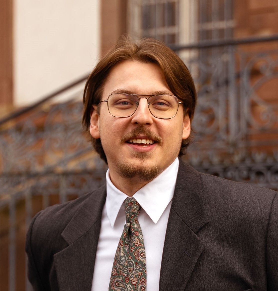 Homme en costume et cravate, souriant à l'extérieur, avec un bâtiment en arrière-plan.