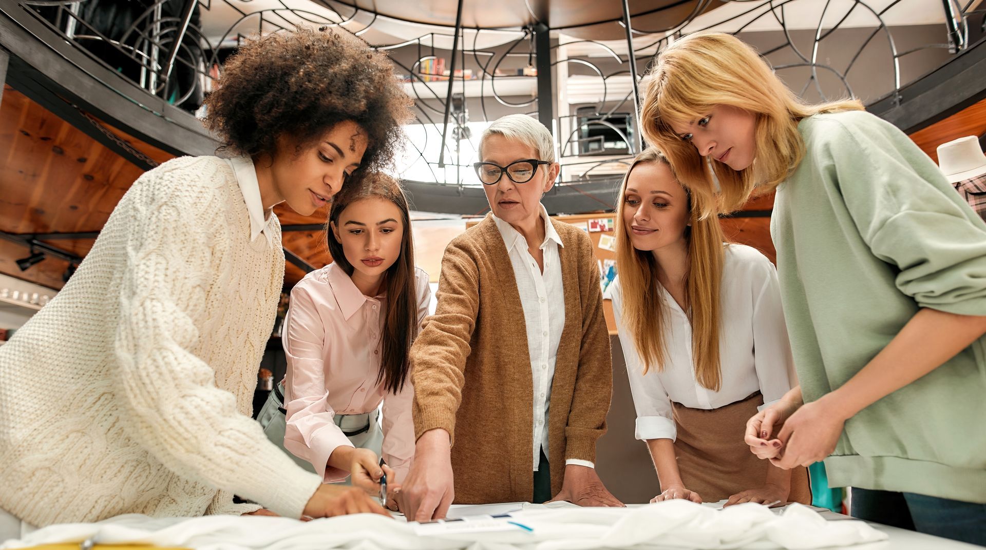Cinq personnes sont réunies autour d'une table et examinent un dessin. Une femme aux cheveux gris dirige le groupe.