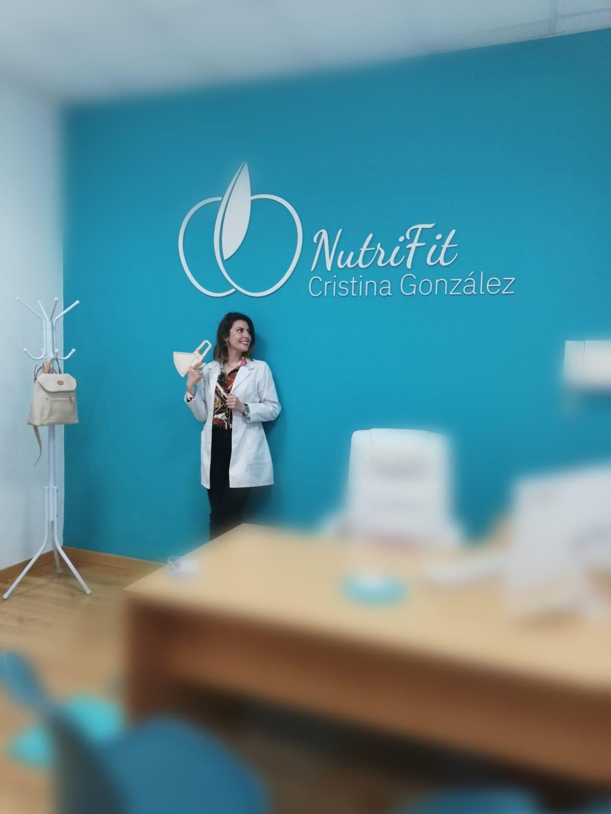 Una mujer con bata blanca está junto a una pared azul con el logotipo de NutriFit.