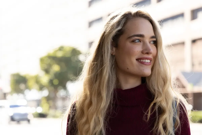 Mujer sonriente con cabello rubio largo y suéter color burdeos al aire libre cerca de edificios.