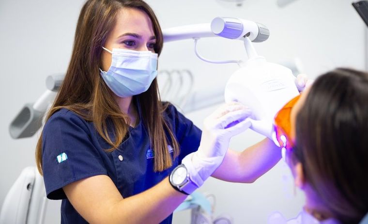 Un odontólogo con bata azul y mascarilla examina a un paciente con una luz dental brillante.
