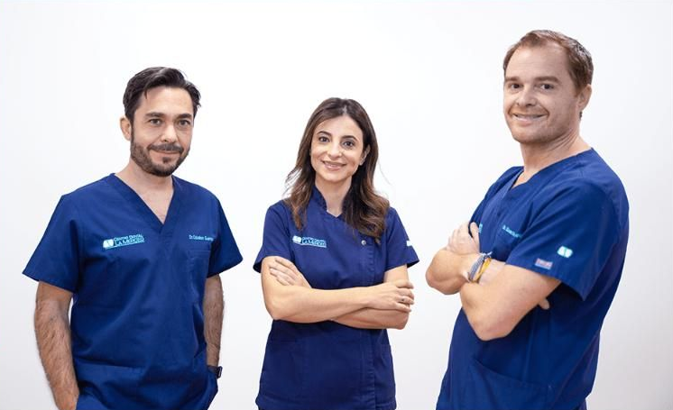 Tres miembros del personal médico con uniformes azules están de pie juntos, sonriendo con los brazos 
