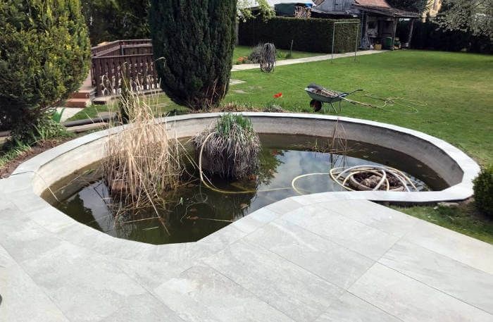 Geschwungener Gartenteich mit Schilf, einem kleinen Springbrunnen und einer Vogelskulptur neben einem Rasen